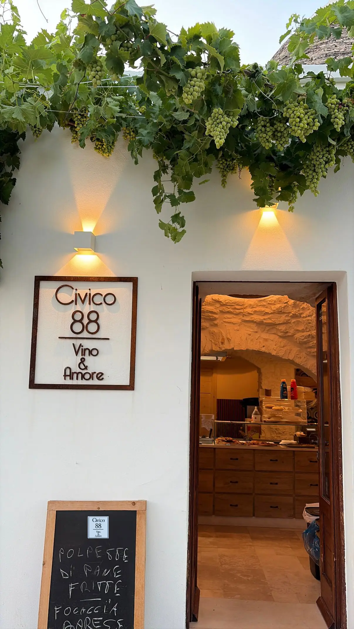 L'ingresso di Civico 88 ad Alberobello: l'insegna in corten sul muro di calce, la vite carica d'uva sopra la porta e il trullo illuminato all'interno